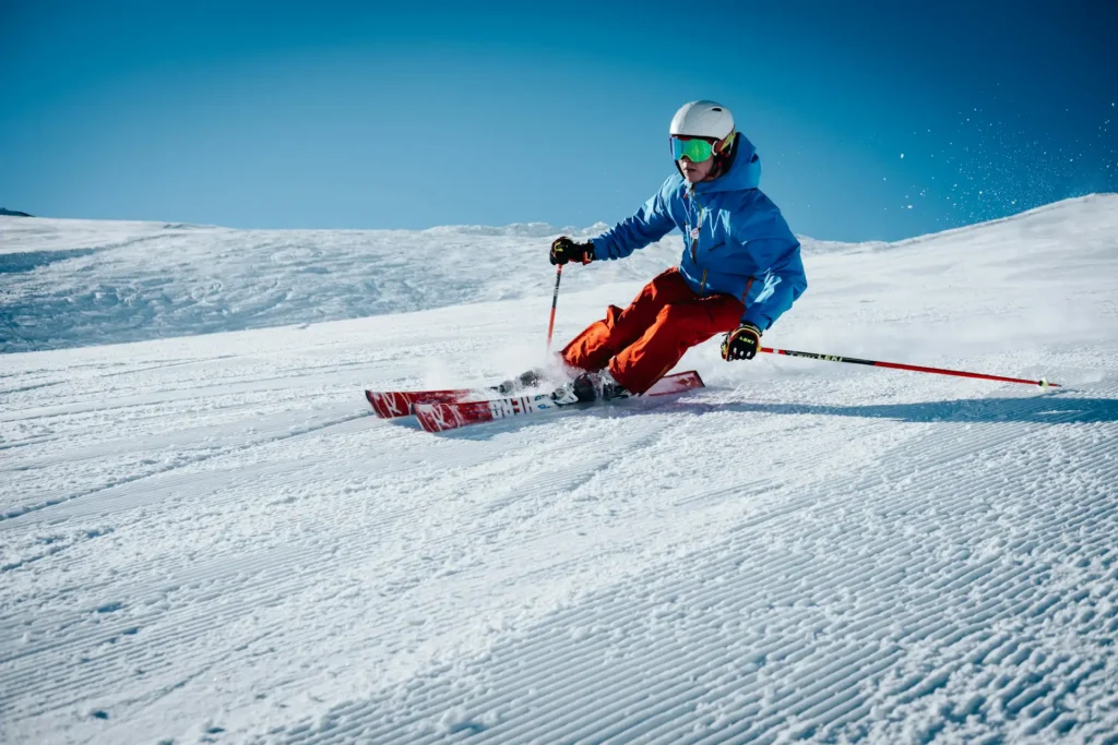 Una persona vestita con tuta da sci con giacca blu e pantaloni rossi pratica sci alpino su una pista. Lo sfondo mostra della neve da poco battuta e cielo azzurro.