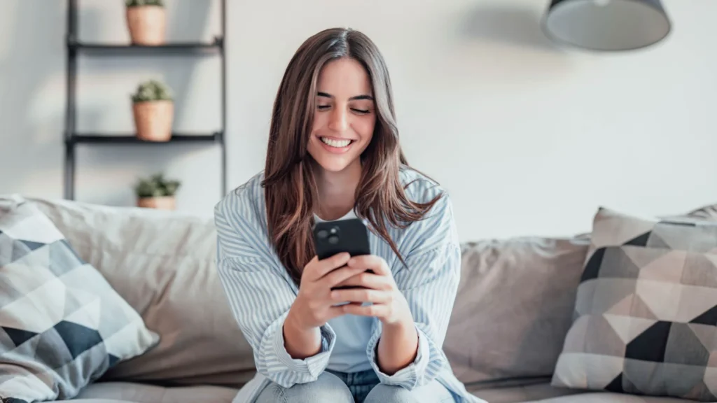 Una ragazza sorridente è seduta su un divano beige e regge tra le mani un cellulare